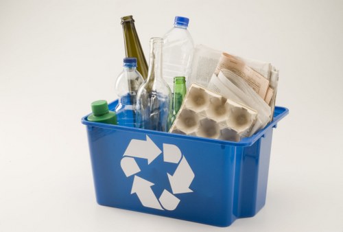 A bright blue plastic recycling bin featuring a white recycling symbol on the front, positioned on a plain white background. The bin is filled with various waste items typical of commercial or office rubbish, including several empty glass bottles in shades of green and brown, with some bottles leaning against each other and slightly protruding from the top rim. There are also transparent plastic bottles, with a visible green cap on one and a blue cap on another, resting among crumpled sheets of paper and newspaper pages. The paper waste appears slightly torn and folded, with visible creases and varying shades of off-white and light grey. The bin's interior is filled to the brim, with some items stacked on top of each other, creating a disorganized appearance. The overall scene suggests a focus on recyclable waste collection, relevant to commercial rubbish removal services provided by Business Waste Removal Clapham, serving the Clapham area of London, with its postcode inferred as SW4.