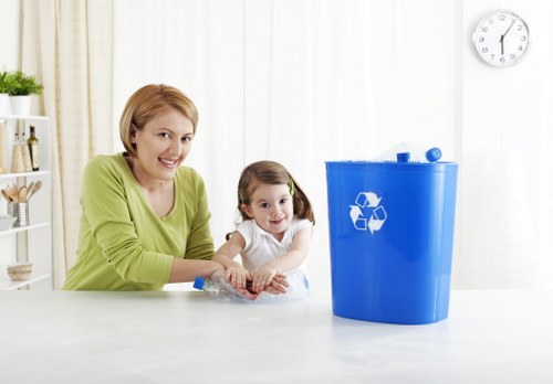A woman with short blonde hair wearing a light green long-sleeved top is seated at a white table during daytime, smiling as she interacts with a young girl who has long brown hair tied in pigtails and is dressed in a white top. The woman’s hands are resting on the table near a clear plastic container, and both are positioned on the left side of the image. To the right, there is a bright blue plastic recycling bin with a white recycling symbol on the front, placed on the white surface of the table. The background features a white wall with vertical blinds, allowing natural light to illuminate the scene, and a small wall clock indicating approximately 12:45. The setting appears to be a clean and modern kitchen or utility area, subtly emphasizing the importance of waste separation, which relates to rubbish and recycling services provided by companies like Business Waste Removal Clapham, operating in the Clapham area.