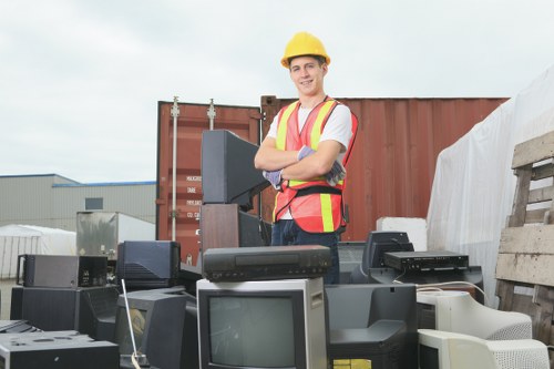 A young male worker wearing a yellow safety helmet and high-visibility vest stands outdoors in an industrial setting, smiling with arms crossed behind a collection of discarded electronic appliances. The appliances include old CRT monitors, microwave ovens, and various computer equipment, arranged on the ground and appearing worn and obsolete. Behind him, there is a large, rusted shipping container and wooden pallets, suggesting a waste disposal or recycling site near Clapham, London. The sky is overcast, providing soft, diffused lighting. The scene reflects a professional rubbish removal operation managed by Business Waste Removal Clapham, highlighting the handling of large bulky waste such as electronic goods, often encountered in commercial clearance projects.