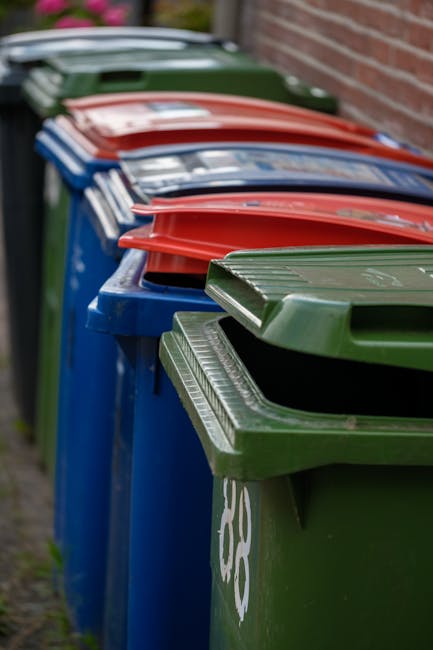 A row of large plastic wheelie bins lined up outdoors along a pavement, with the front green bin in clear focus and the others extending into the background. The bins are predominantly blue, green, re
